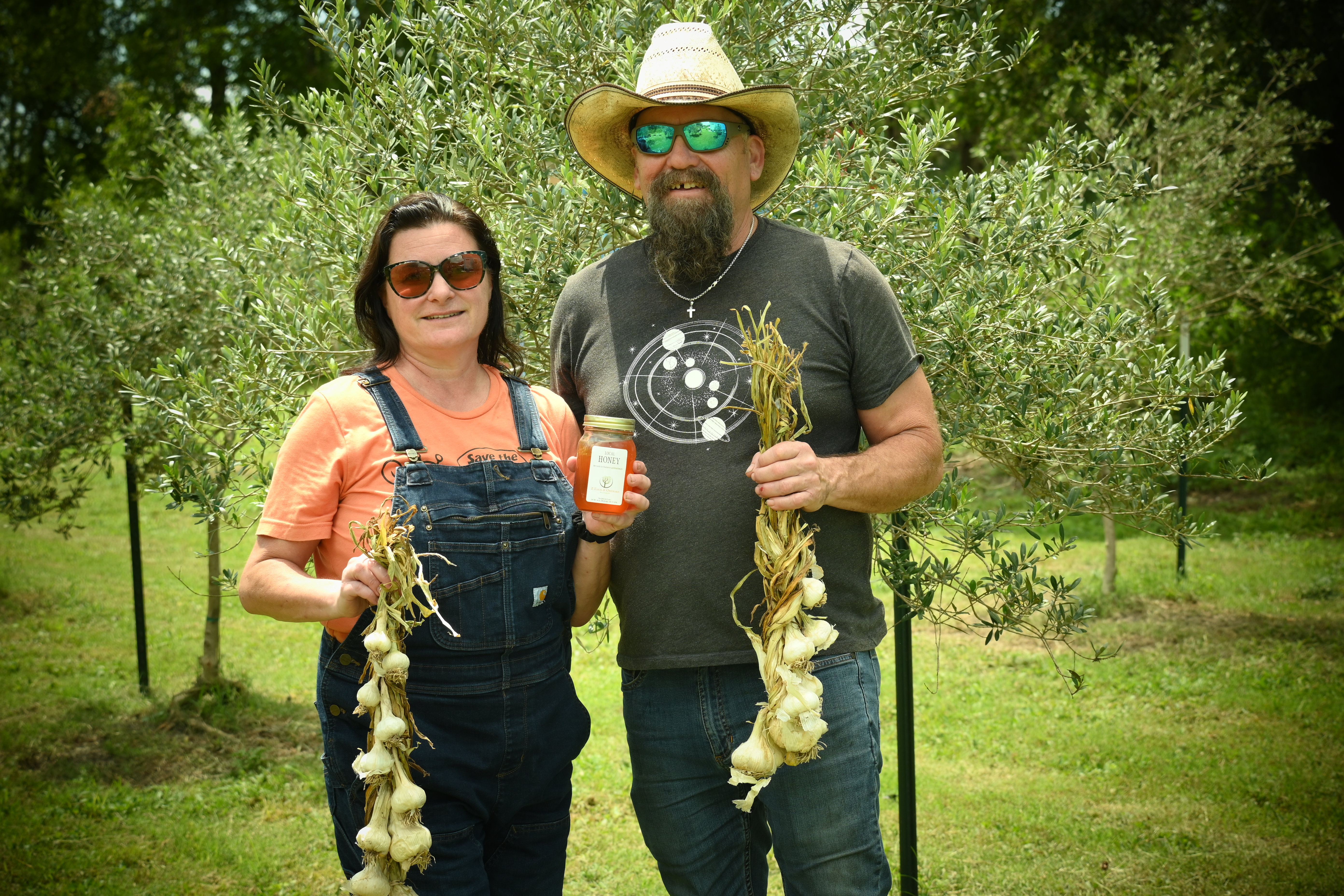 A man and a woman pose in front of trees and hold garlic and jars.